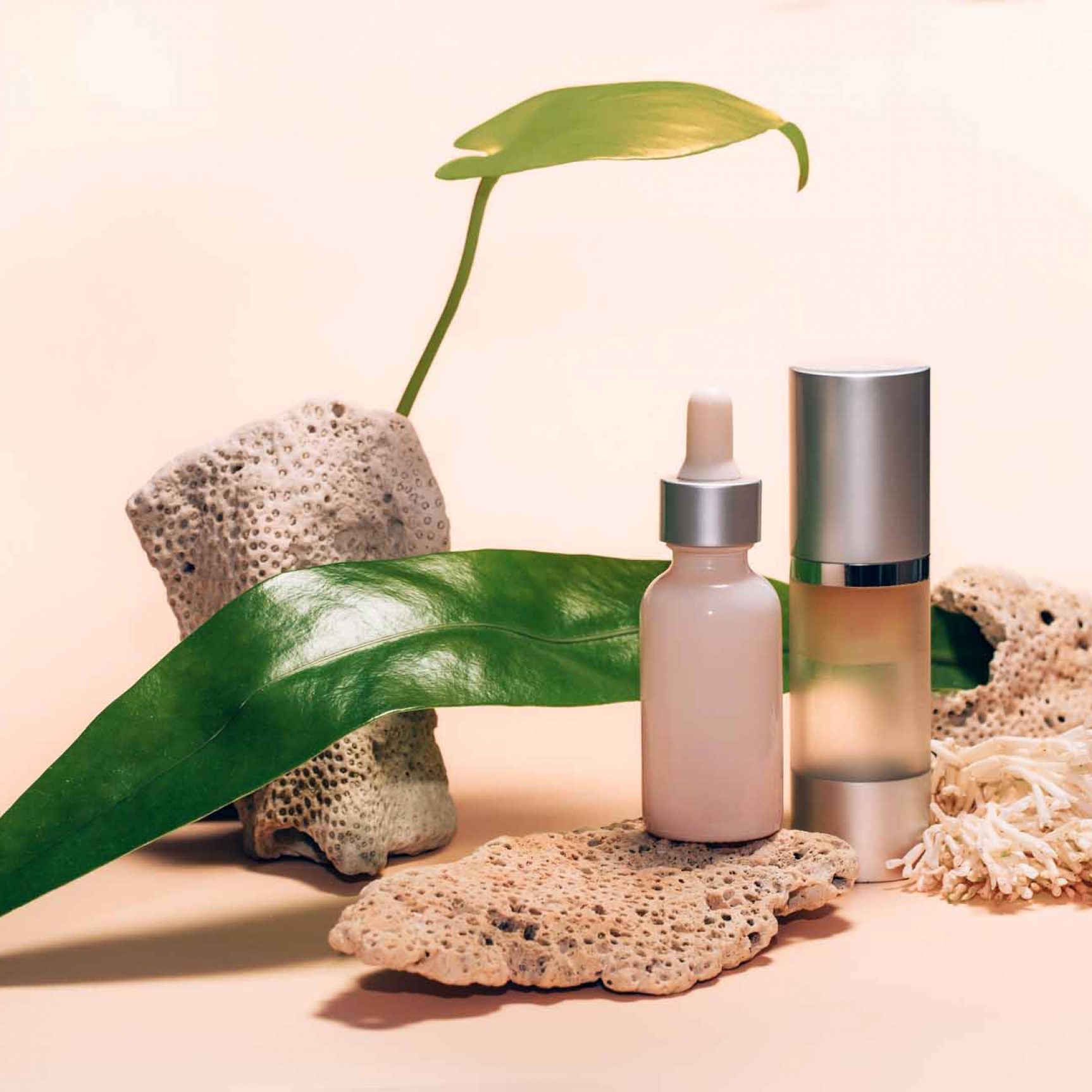 Decorative composition with cosmetics, woman beauty products. Droplet and pump bottles with serum and oil with corals and green leaves on a beige background.